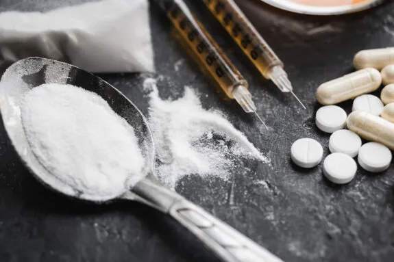 Narcotic substances in spoon, dope powder in transparent plastic bag, syringes with drugs dose, white pills on dark textured background.